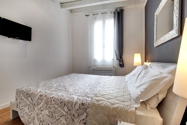 A bedroom with a bed, a window with curtains, an air conditioning unit, a wall-mounted mirror, and a bedside lamp.