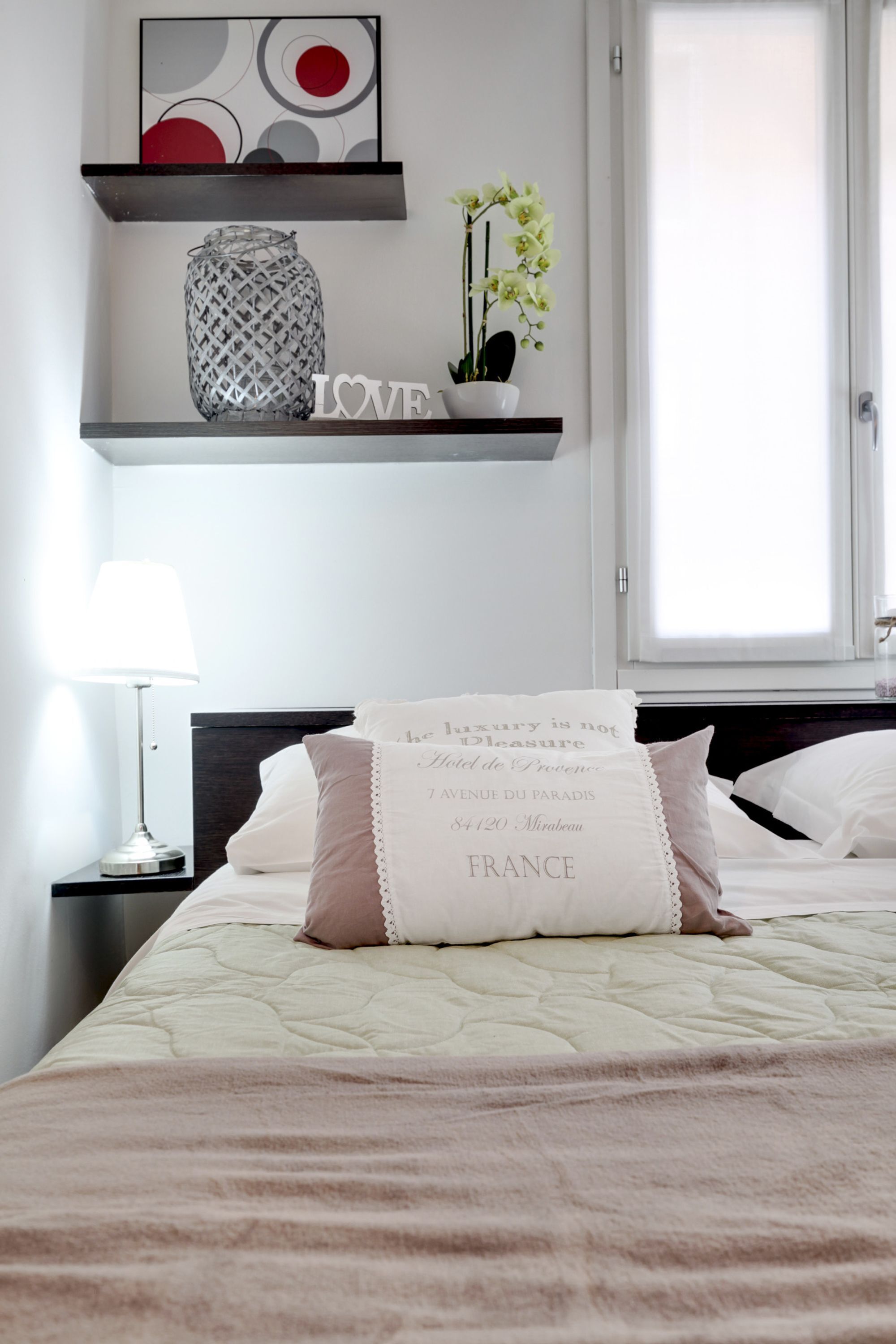 Un lit bien fait, recouvert d’une courtepointe matelassée et agrémenté de coussins décoratifs, une lampe de chevet, une étagère avec un vase et des fleurs, et une œuvre d’art encadrée au-dessus du lit.
