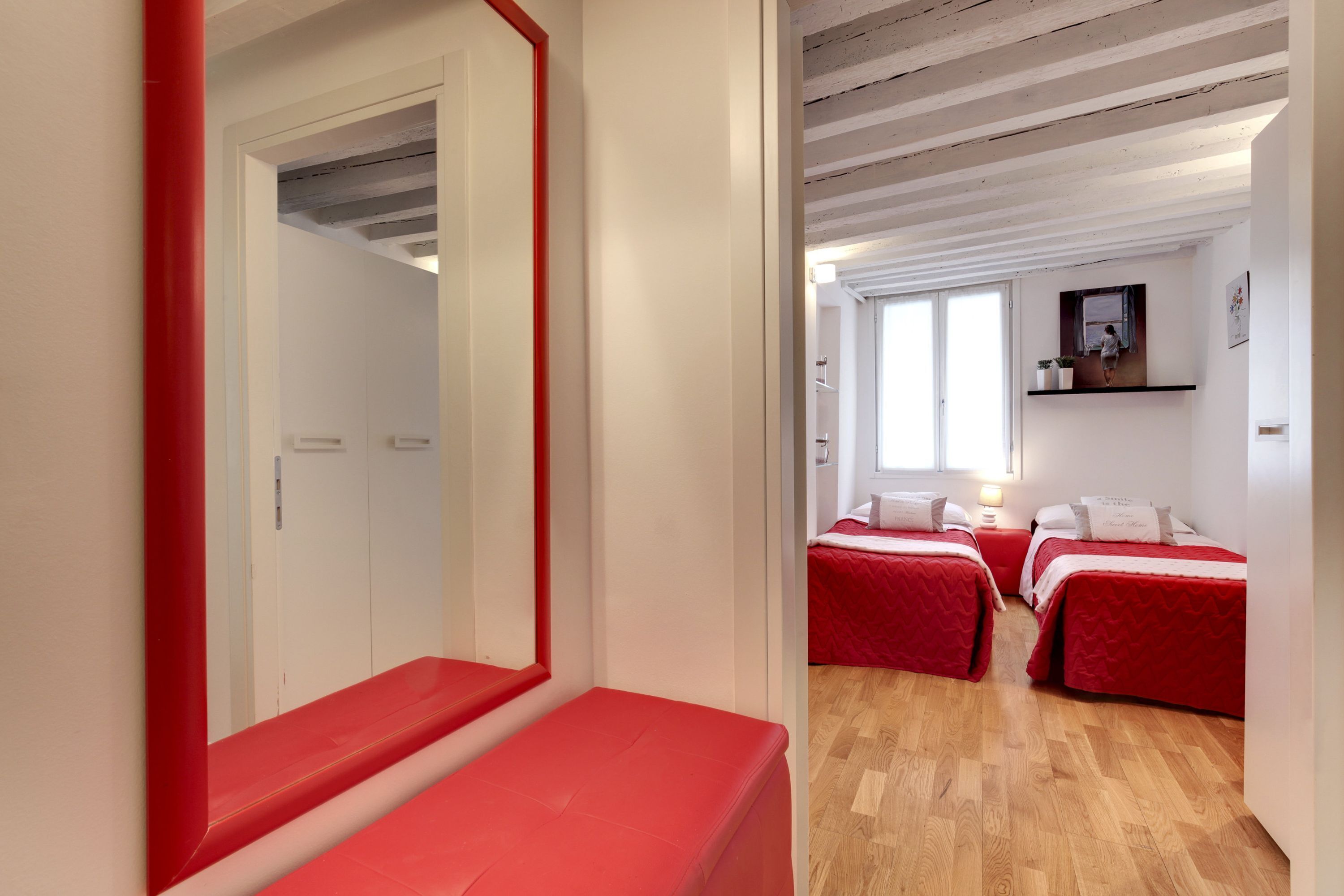 Quarto com três camas, piso de madeira e um banco vermelho.