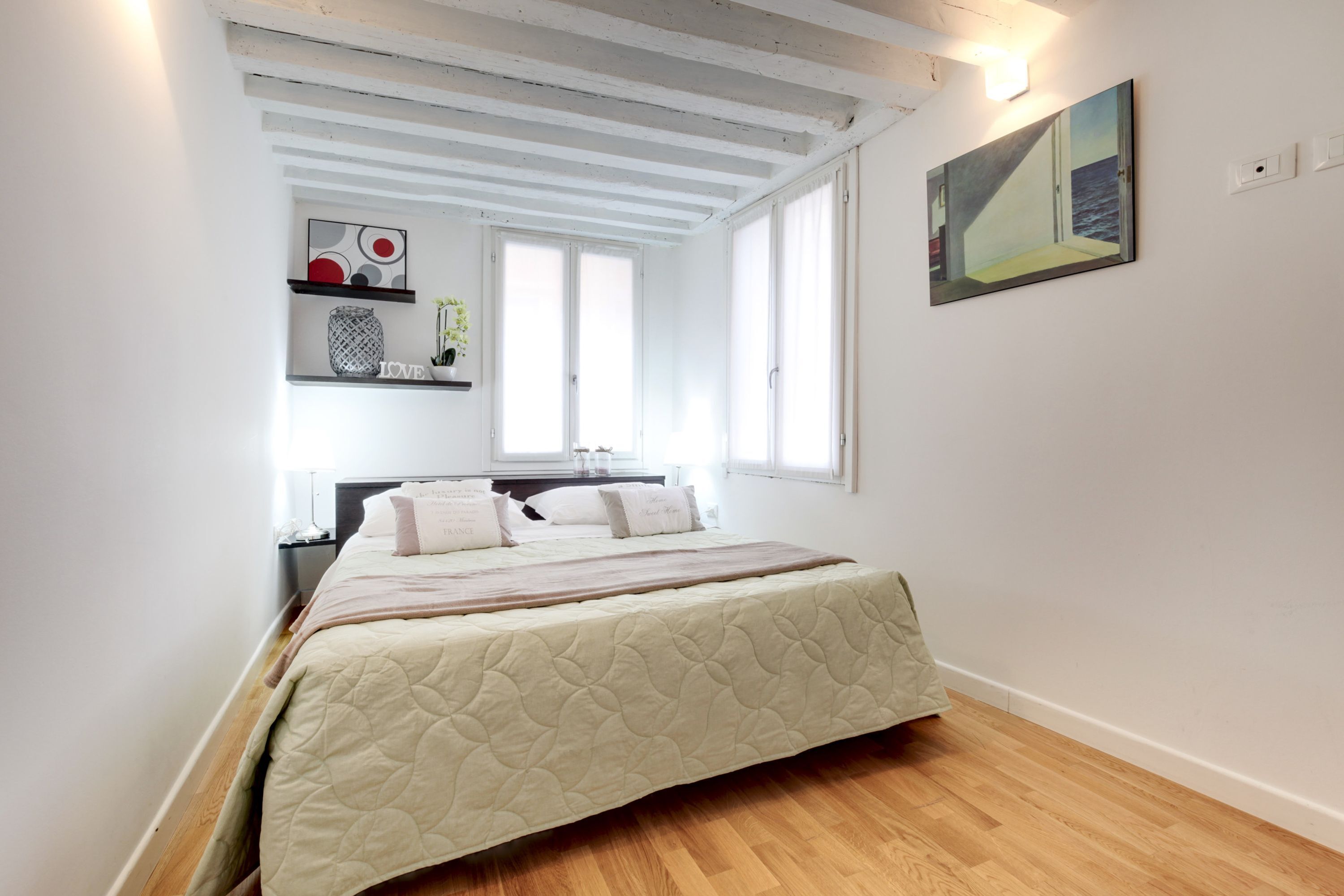 A bedroom with a bed, a shelf with decorative items, a painting, and a window.