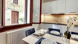Ett modernt matområde med ett bord dukat för två personer, utsikt över en byggnad i sten och ett kök med vita skåp.