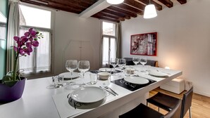 Une salle à manger avec une longue table dressée pour six personnes, des assiettes blanches, des verres à vin et des couverts. Un vase rempli de fleurs violettes se trouve sur la gauche, et un tableau est accroché au mur.