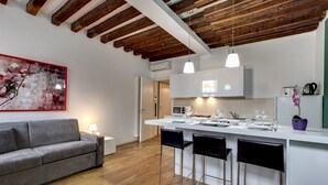 Une cuisine moderne avec des meubles blancs, un îlot central doté de tabourets de bar et un canapé gris dans un salon lumineux.