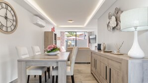 Un comedor moderno con mesa de madera y sillas blancas, una cocina con mueble de color claro y un reloj colgado en la pared.
