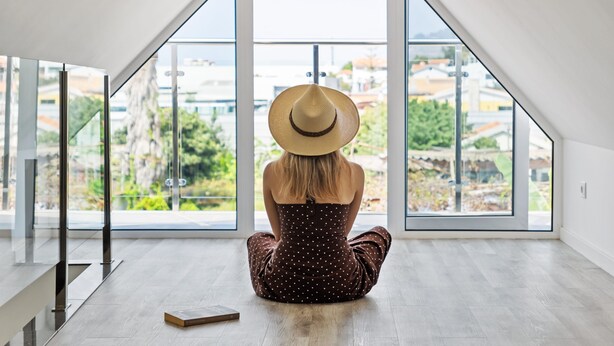 Una persona sentada en el suelo de una habitación moderna con ventanas amplias, piso de madera y barandilla de vidrio.
