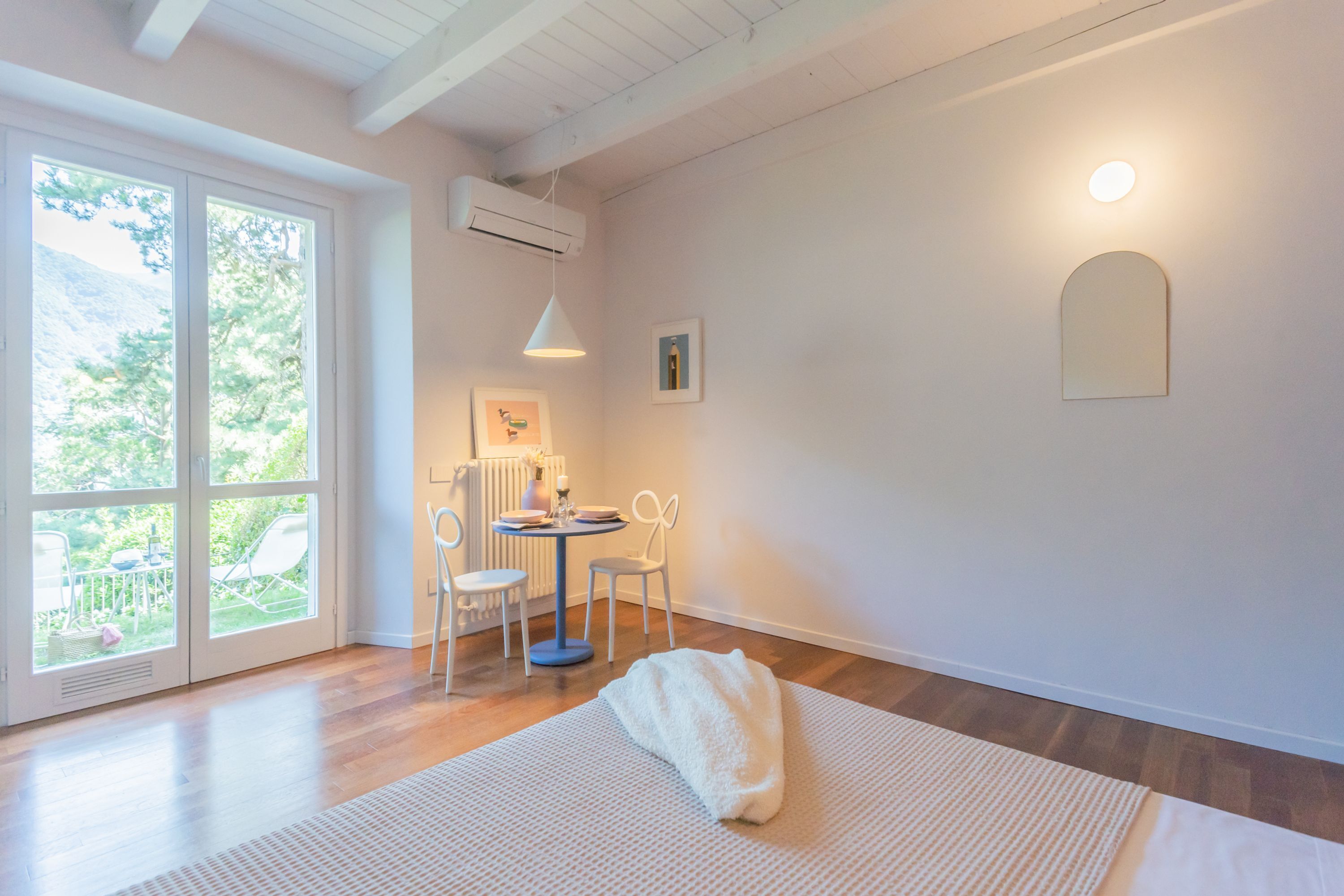 Une pièce lumineuse avec un plancher en bois, un canapé blanc, une table avec des chaises et une grande fenêtre donnant sur un paysage verdoyant.