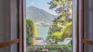 A view from a room with a glass door, overlooking a lake and mountains, with two chairs and a table set outside. - OC Lake Love in Laglio (Laglio)