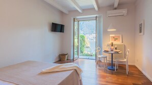 A bedroom with a bed, a TV, a dining area with a table and chairs, and a view of a mountain through a glass door. - OC Lake Love in Laglio (Laglio)
