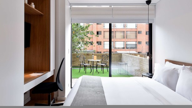 A hotel room with a bed, a desk, a chair, a small table, and a view of a brick building and greenery outside. - w Radiant Loft w Amazing Terrace in Bogota (Bogotá)