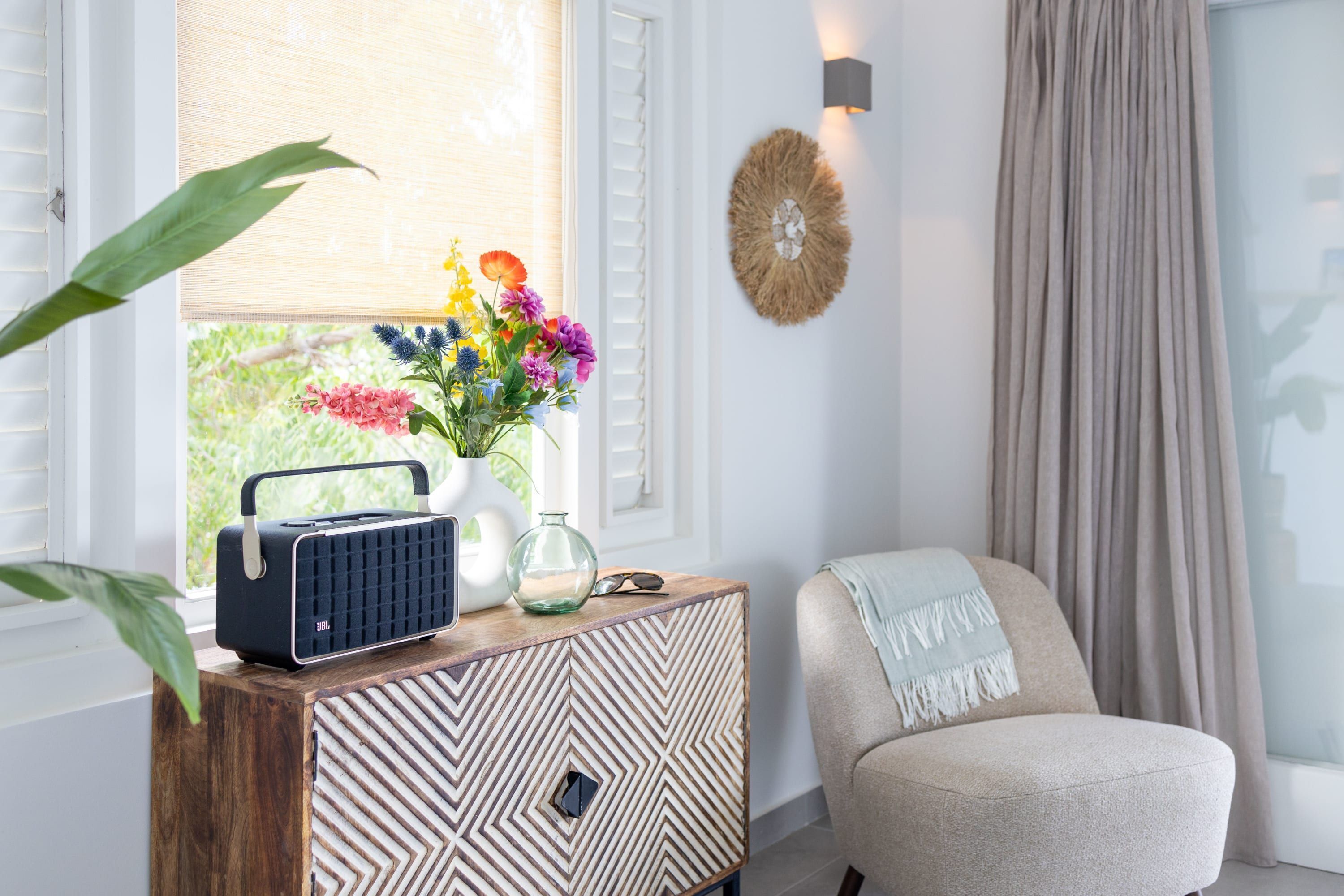 A room with a wooden sideboard, a chair, a window with blinds, a wall-mounted light, and a decorative wall piece.