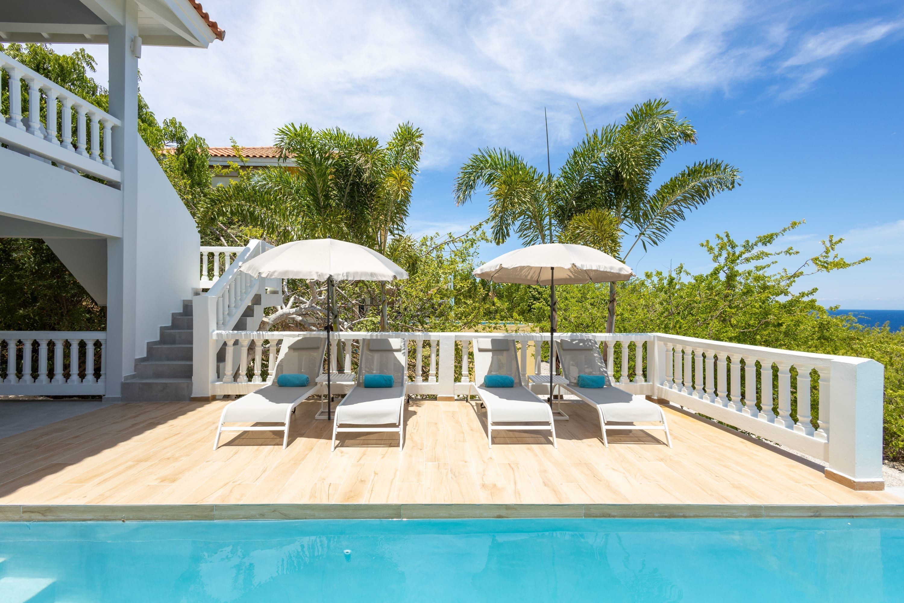 A poolside area with white lounge chairs and umbrellas, a wooden deck, and a view of the ocean.