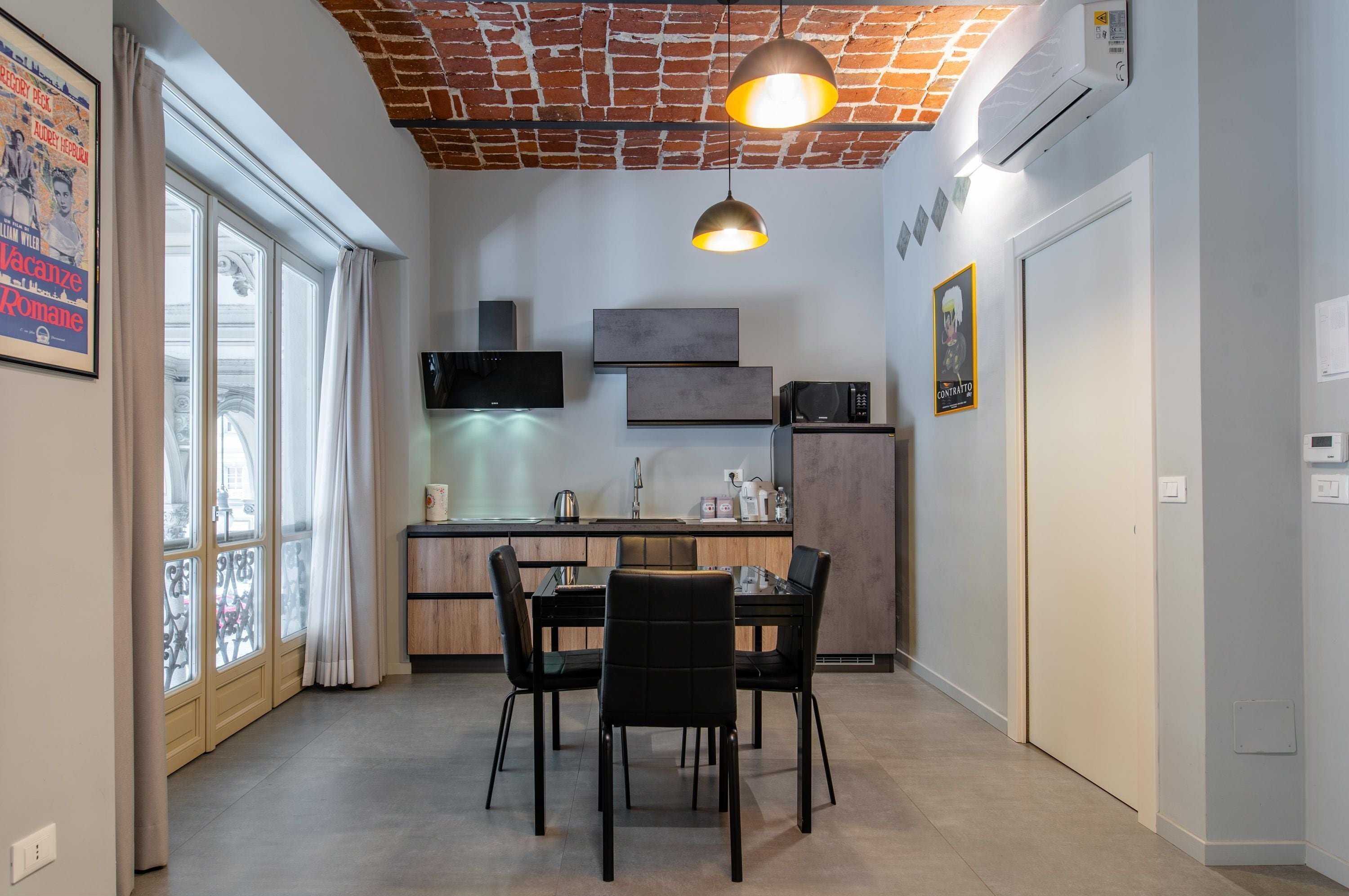 A modern kitchen with a dining area, featuring a brick ceiling, pendant lighting, and a large window with curtains.