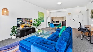 Un moderno salón con un sofá azul, una mesa de centro de vidrio y un televisor de pantalla plana. Hay una zona de comedor con una mesa y sillas, y una cocina al fondo.