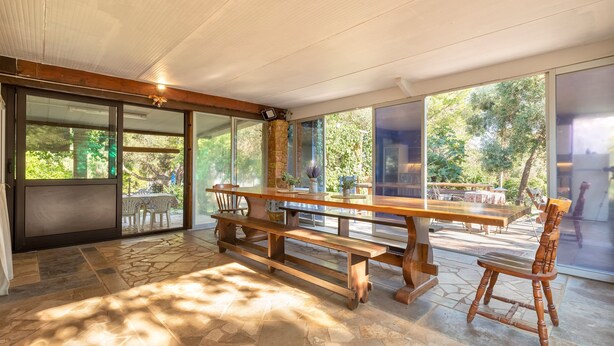 An open-plan living area with a wooden bench and table, large windows, and a stone floor.
