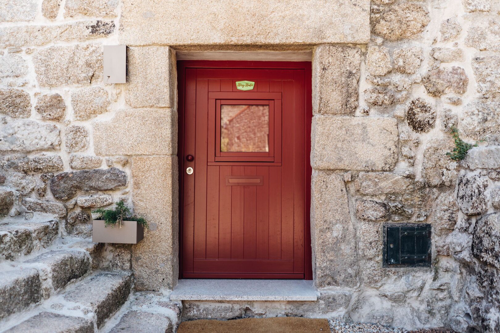 Una porta rossa con un piccolo oblò sopra, incastonata in un muro di pietra.