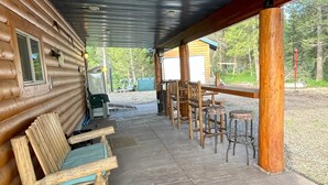 Een overdekt terras met houten banken en tafels, een overdekte doorgang en een uitzicht op een houten chalet en bomen op de achtergrond.