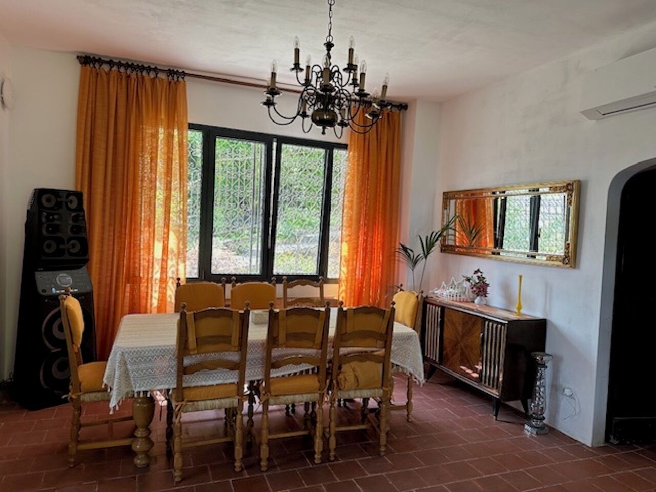 A dining area with a table set for four, surrounded by wooden chairs. A chandelier hangs from the ceiling, and there is a radiator on the wall.
