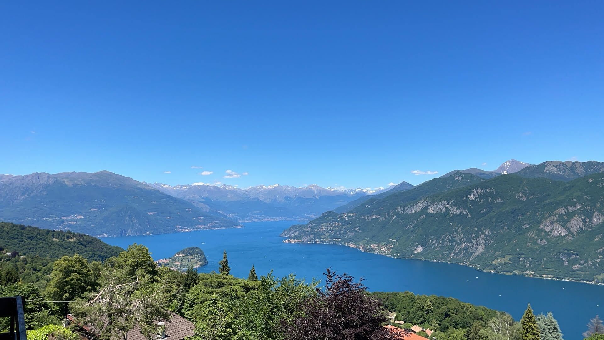 A panoramic view of a mountainous landscape with a lake, surrounded by dense forests and distant snow-capped peaks.