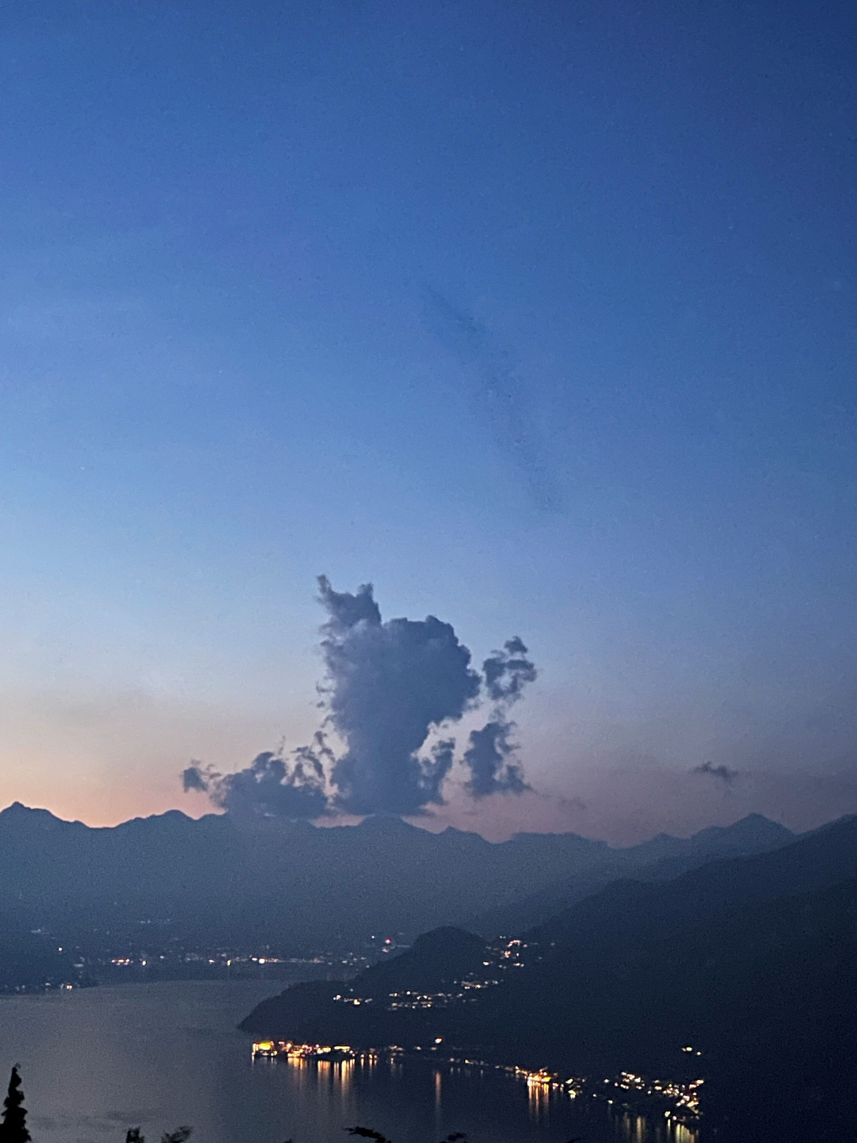 Une vue nocturne d’un lac, avec des montagnes en arrière-plan et une silhouette urbaine dont les lumières se reflètent sur l’eau.