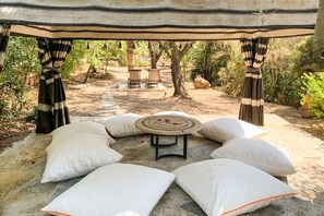 An outdoor seating area with a round table and white cushions, surrounded by trees and a stone pathway.