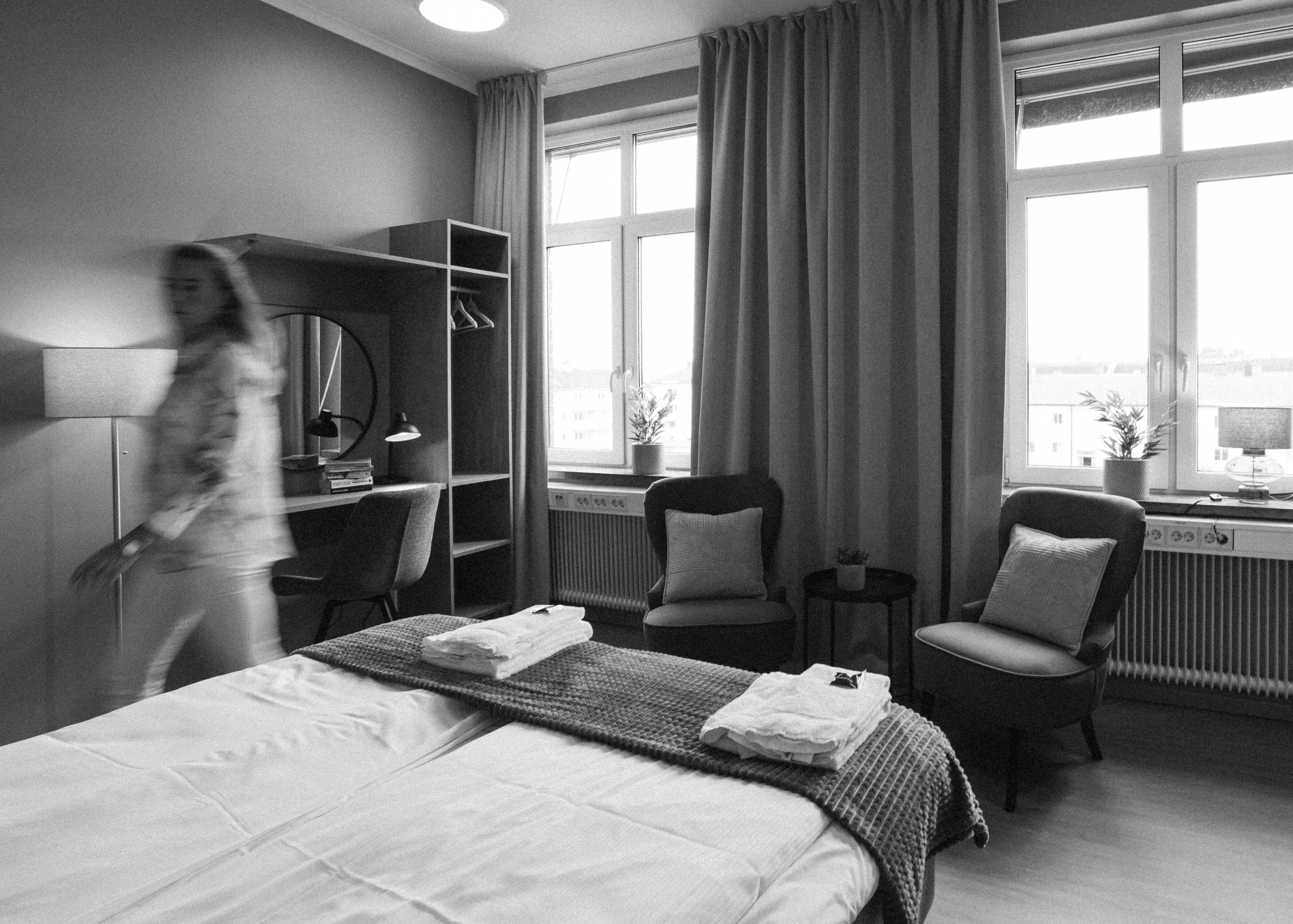 Habitación de hotel con cama, dos sillas, escritorio, armario y ventanas amplias.