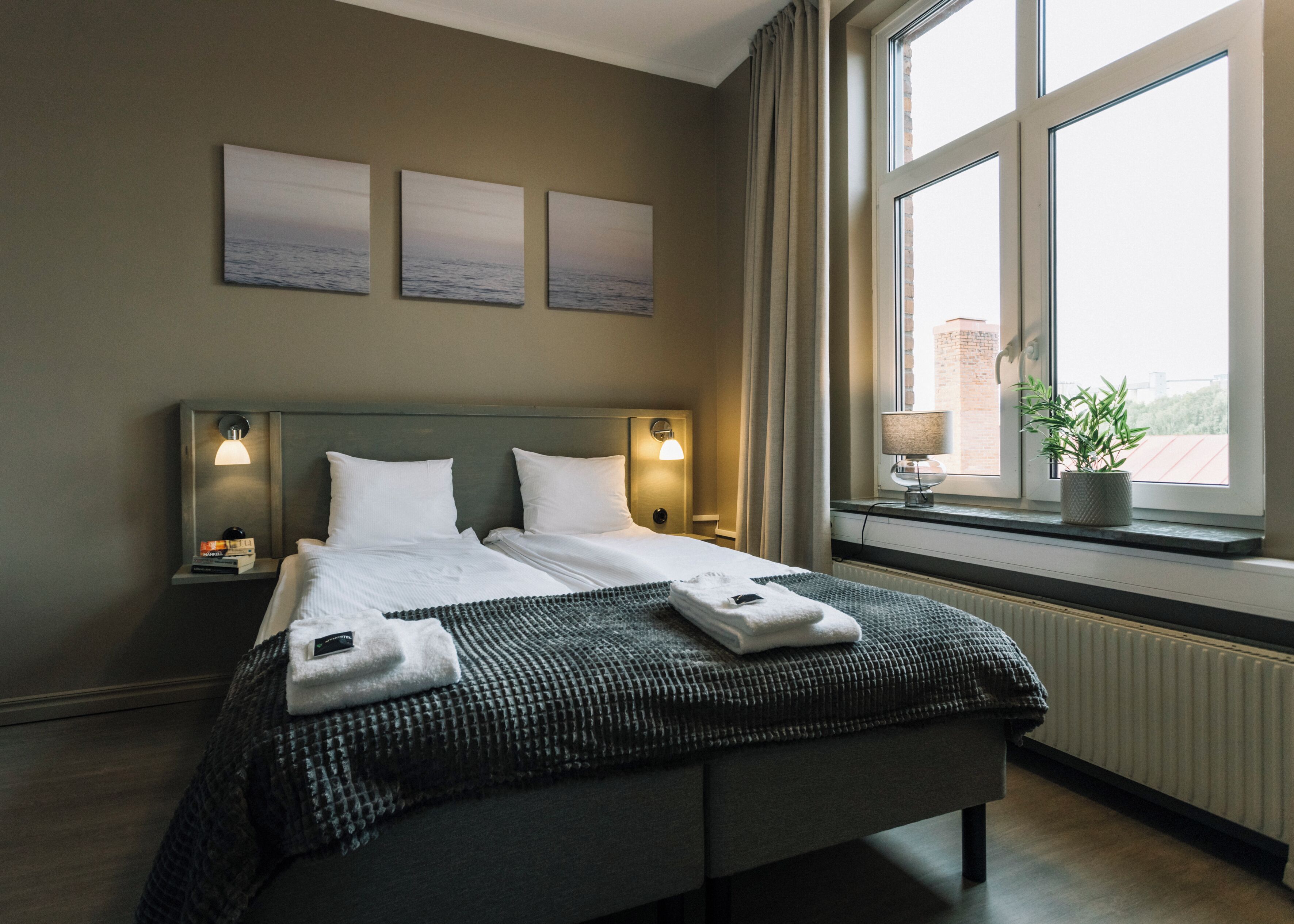 Una cama bien hecha con almohadas y una colcha, una mesita de noche con una lámpara y una ventana con vistas a edificios.
