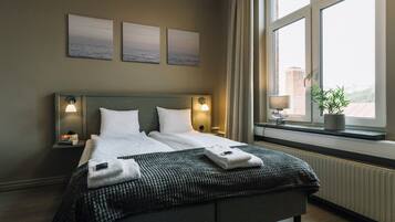 Una cama bien hecha con almohadas y una colcha, una mesita de noche con una lámpara y una ventana con vistas a edificios.