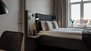 A modern hotel room with a large bed, a bedside table with a bottle and glasses, a chair, and a window with a view of buildings. - My Hostel Lidköping (Lidköping)