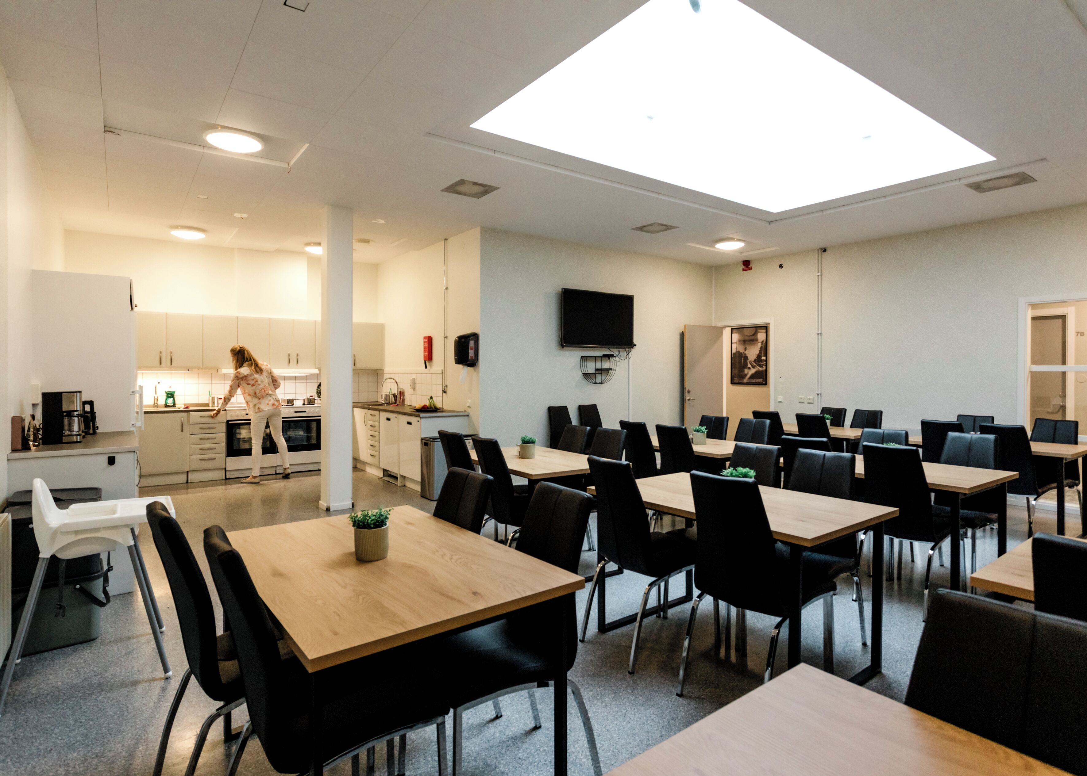 A modern dining area with multiple tables and chairs, a kitchen in the background, and a flat-screen TV mounted on the wall.