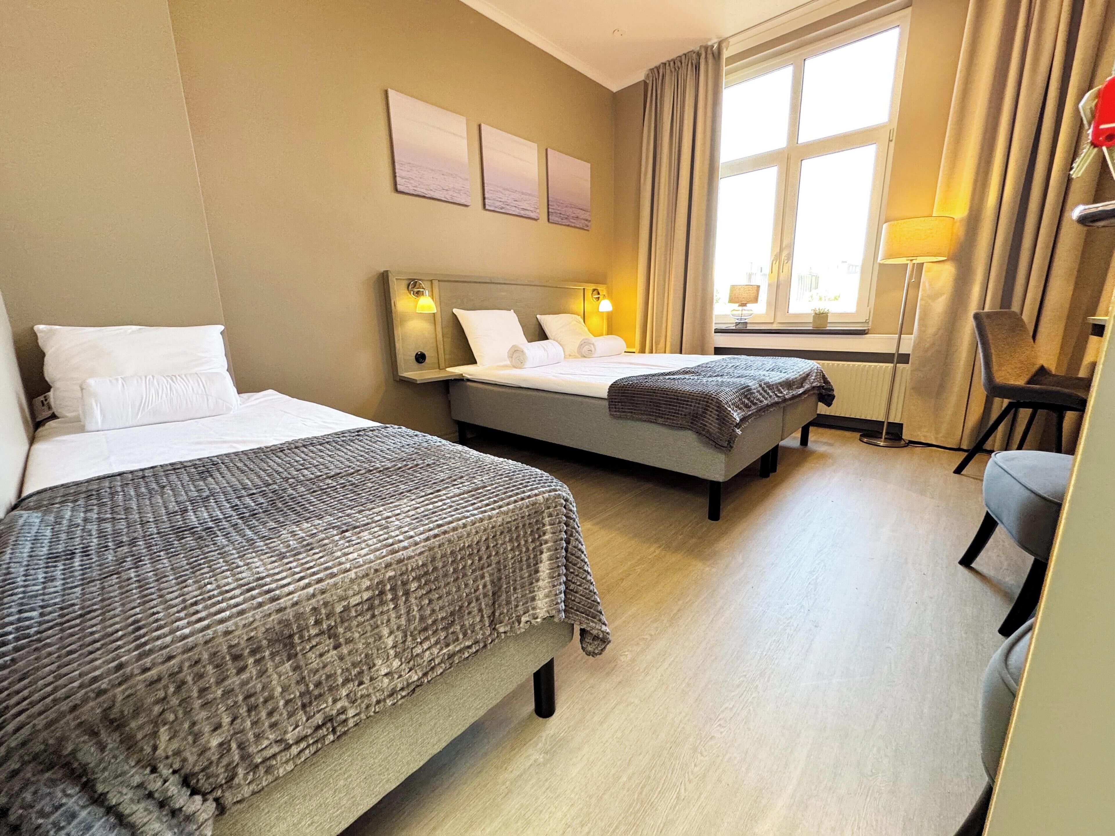 A hotel room with two beds, a desk, and a chair. There are three wall-mounted lamps and a window with curtains.