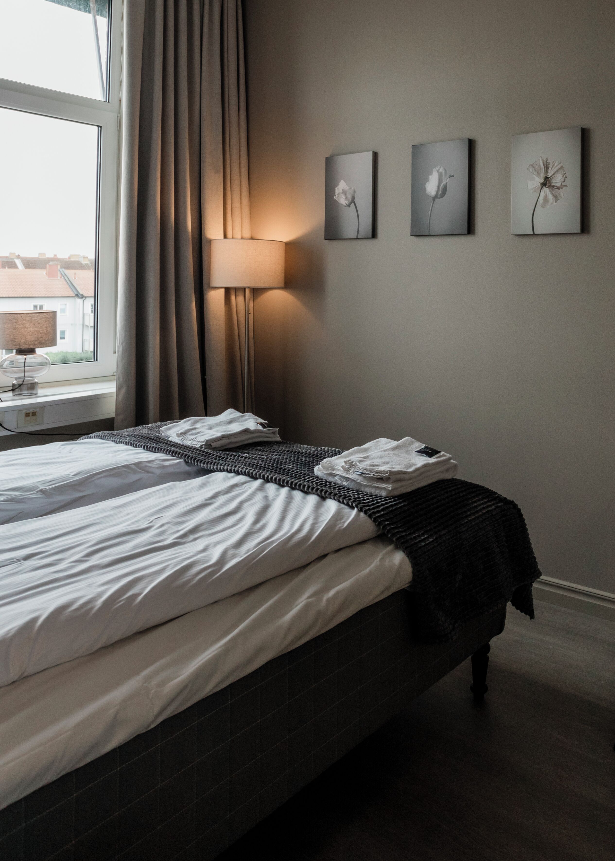 A neatly made bed with white linens and a dark headboard, a bedside lamp, and three wall-mounted artworks depicting flowers.