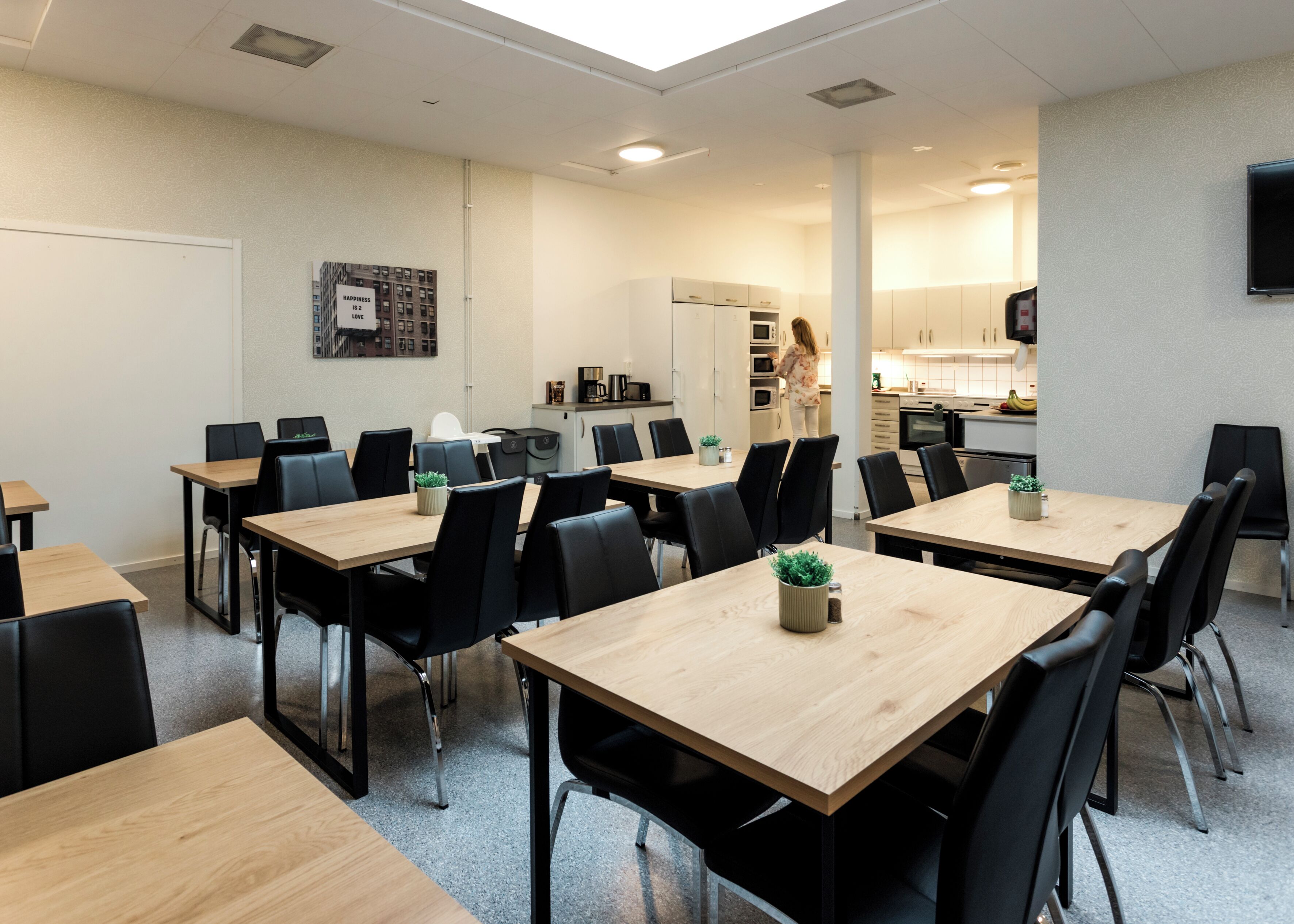 Una sala de reuniones moderna con varias mesas y sillas, una kitchenette al fondo y un televisor montado en la pared.