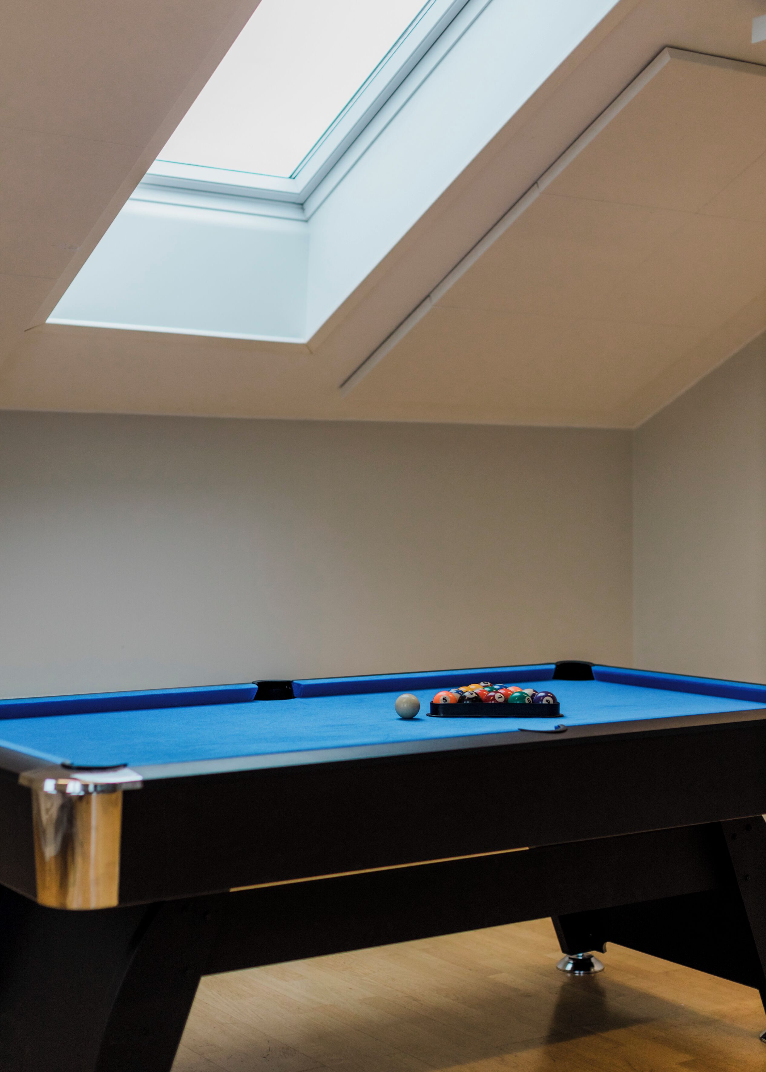 Une table de billard recouverte d’un feutre bleu, un plafond blanc avec une lucarne et un plancher en bois.