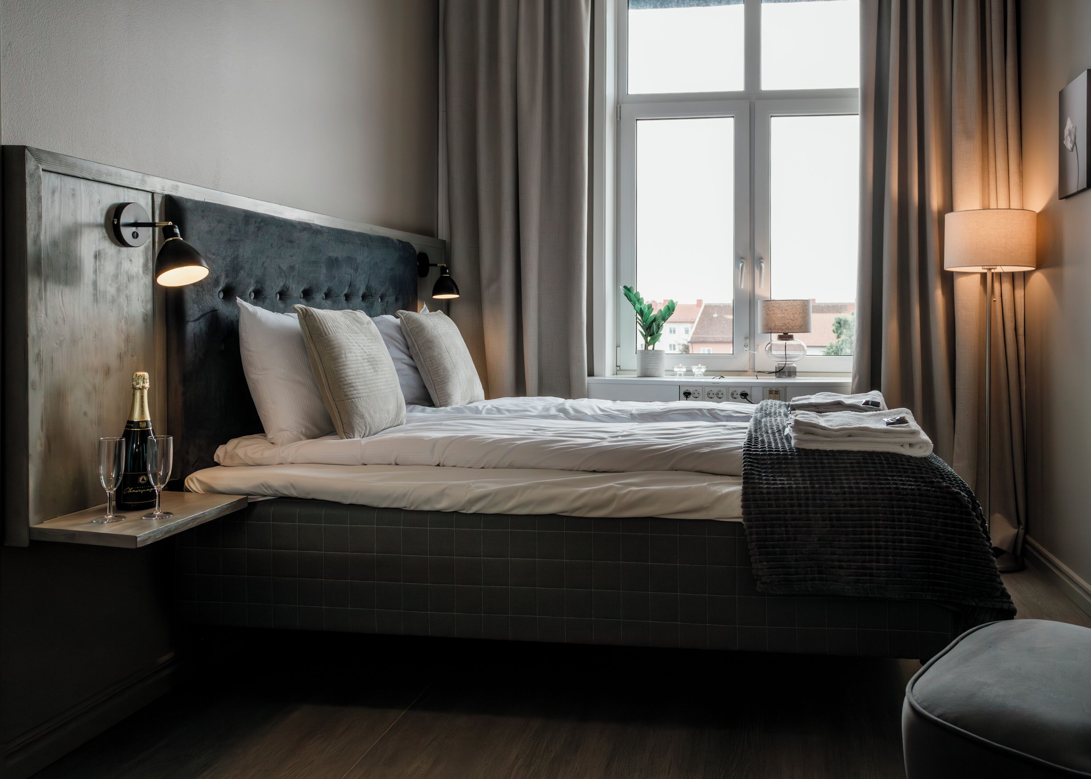 Quarto de hotel moderno com uma cama grande, cabeceira de madeira, mesa de cabeceira com garrafa e copos, janela com cortinas e uma luminária.