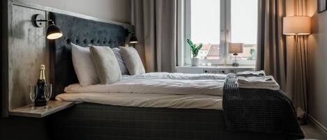 Quarto de hotel moderno com uma cama grande, cabeceira de madeira, mesa de cabeceira com garrafa e copos, janela com cortinas e uma luminária.