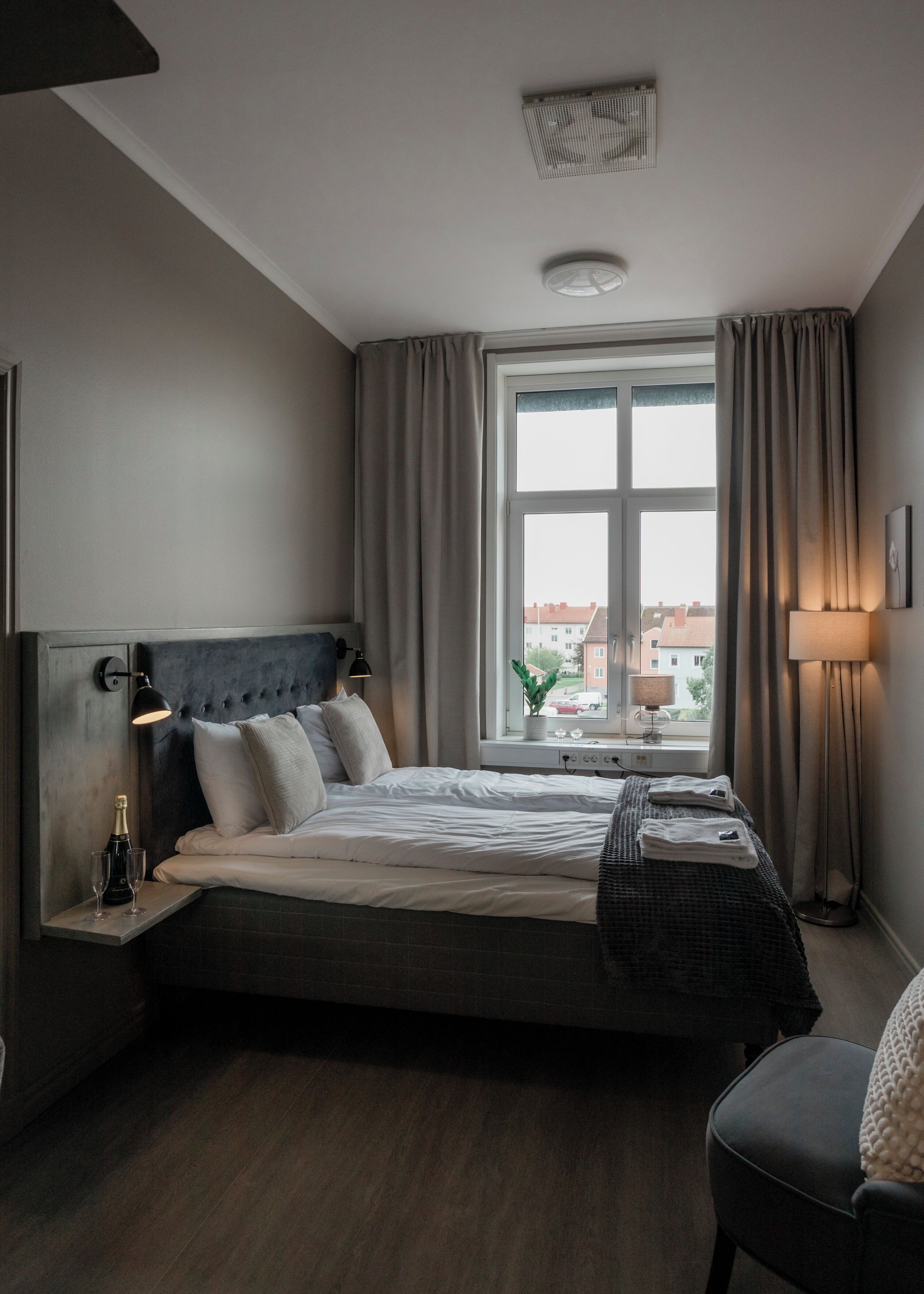 Une chambre d’hôtel moderne avec un grand lit, une table de chevet avec une bouteille et des verres, une fenêtre avec des rideaux et une vue sur les bâtiments environnants.