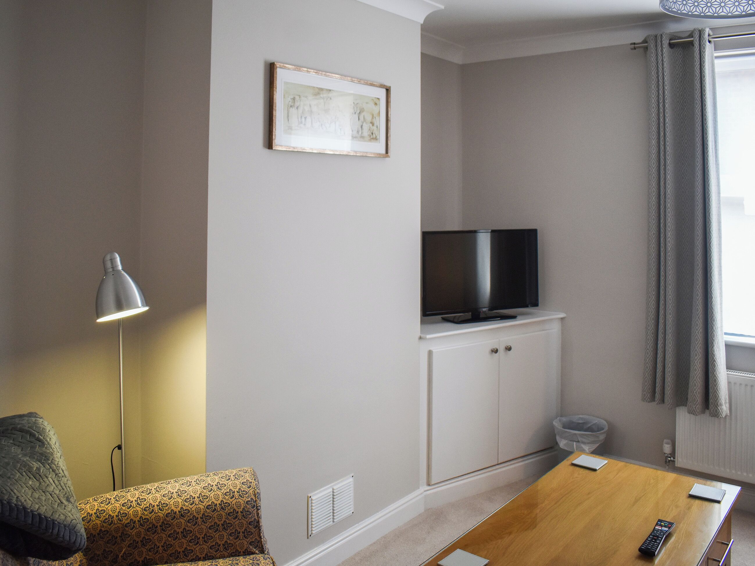 A room with a sofa, a TV on a cabinet, a lamp, a table with a remote, and a window with curtains.
