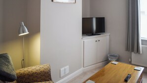 Una habitación con sofá, televisión sobre un mueble, lámpara, mesa con control remoto y ventana con cortinas.