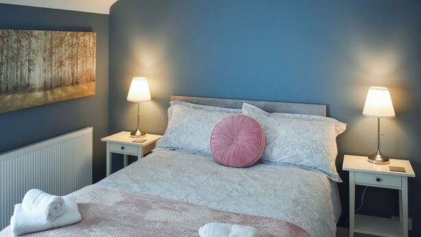 Un dormitorio con una cama, dos mesitas de noche, un radiador, un cuadro en la pared y dos lámparas.