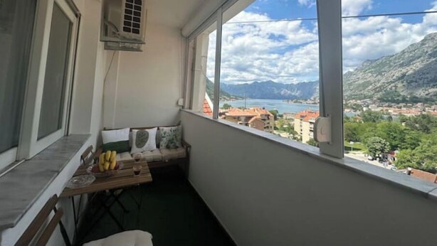 A balcony with a view of a lake and mountains, a small table with fruits, and a chair.
