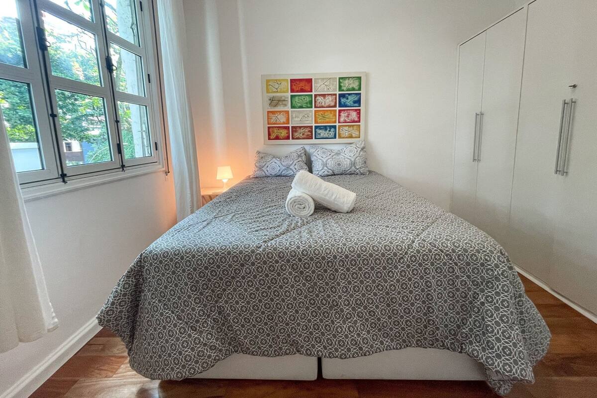 Uma cama bem arrumada com roupa de cama estampada, uma parede com obras de arte coloridas e uma janela com cortinas.