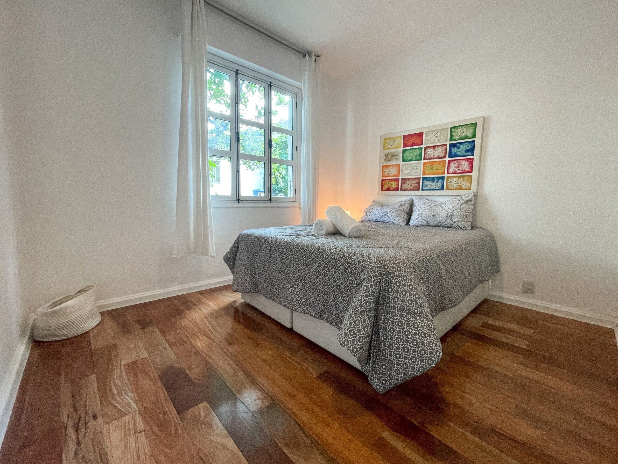 A bedroom with a bed, a window, and a colorful artwork on the wall.