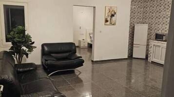 Ein geräumiger Wohnbereich mit einem schwarzen Ledersofa, einem schwarzen Sessel und einer Topfpflanze. Der Raum hat einen gefliesten Boden und eine weiße Tür zu einem anderen Zimmer.
