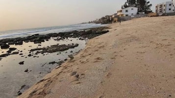Ein sandiger Strand mit vereinzelten Felsen und Fußabdrücken, ein ruhiges Meer und Gebäude im Hintergrund.