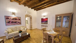 A living room with a wooden floor, a sofa, a coffee table, and a dining table with chairs. There are paintings on the walls and a cabinet with glass doors. - Cagliari Homes (Cagliari)