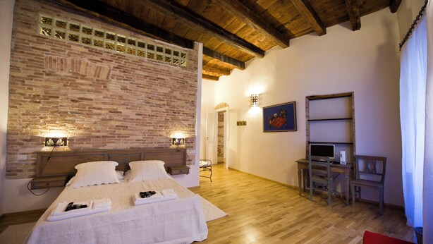 A bedroom with a brick wall, wooden ceiling, a bed with white linens, a desk with a computer, and a chair. - Cagliari Homes (Cagliari)