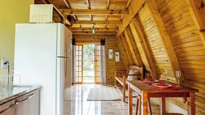 A kitchen with a refrigerator, wooden cabinets, a dining table set for two, and a view of the outdoors through a door. - Chale 2 Familia (Angra dos Reis)