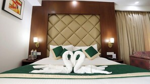 A hotel room with a large bed, a headboard with a diamond pattern, two wall-mounted lamps, a green and beige bedspread, and a heart-shaped towel arrangement on the bed. - Comfort Hotel GMS Grand  (Dehradun)