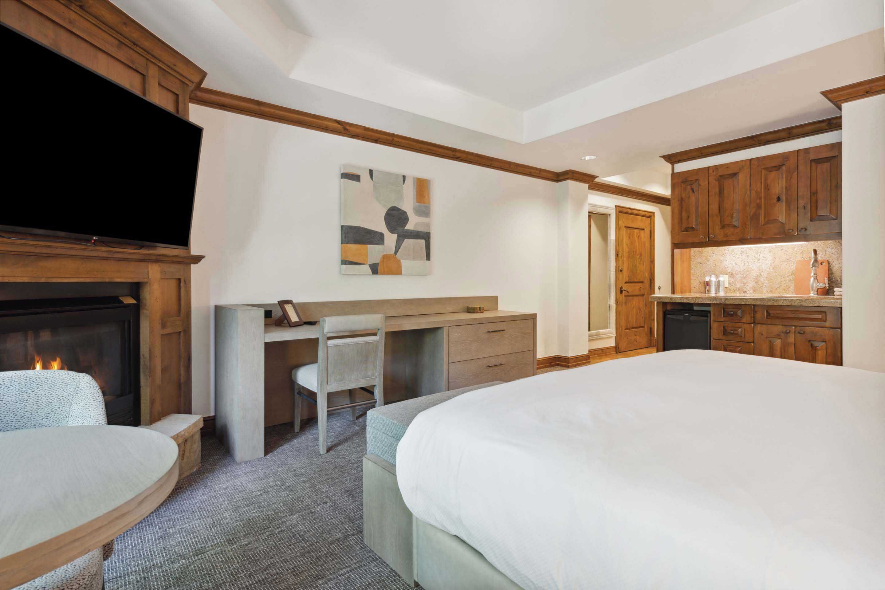 A hotel room with a bed, a desk, a chair, a fireplace, and a kitchen area.