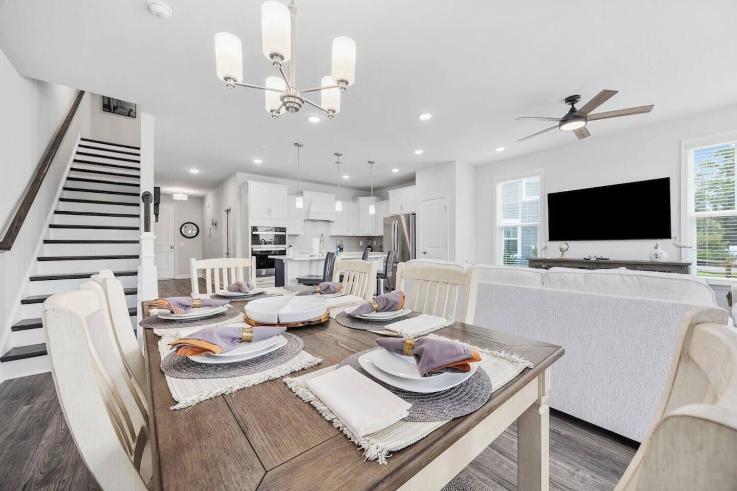 Una cocina moderna con armarios blancos, un comedor con mesa de madera dispuesta para cuatro comensales y una escalera con barandillas oscuras.